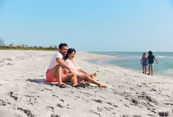 A couple sitting on top of a sandy beach with children in background