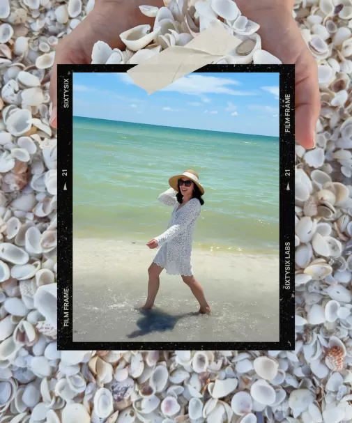Black film-frame overlay taped onto a bed of small white shells, featuring a smiling woman in a straw hat and white crochet cover-up wading along the shoreline.
